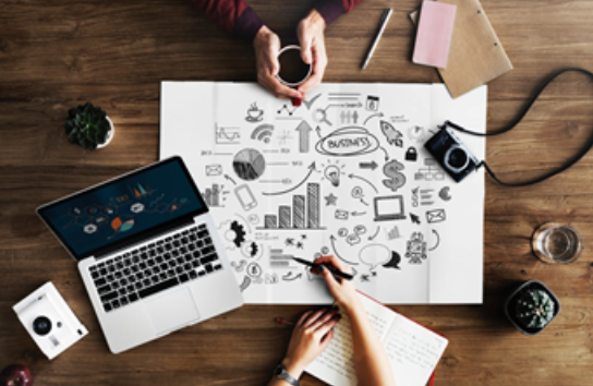 A desk with a computer, drawing of business plan, and hands working.