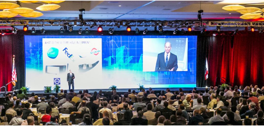 Auditorium at general contractor trade show with full crowd and large video screen