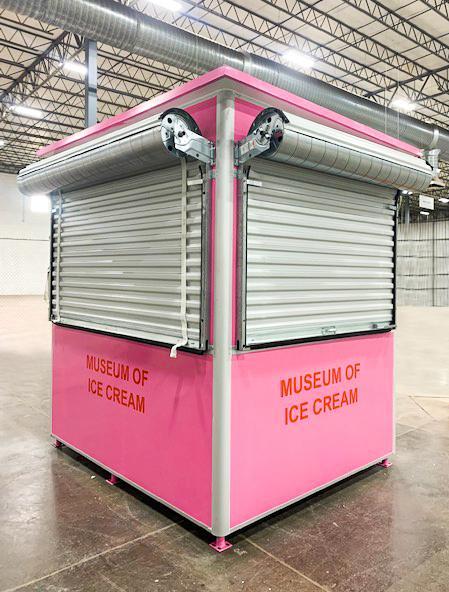 A bright pink modular structure inside of a large room, with closed metal roll-up windows and the words ‘Museum of Ice Cream’ printed in red