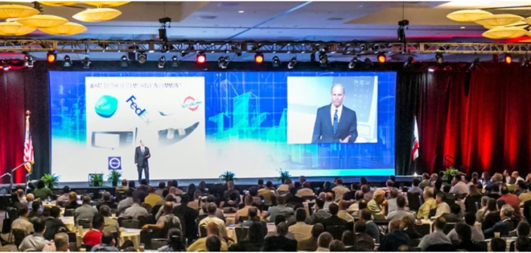 Auditorium at general contractor trade show with full crowd and large video screen